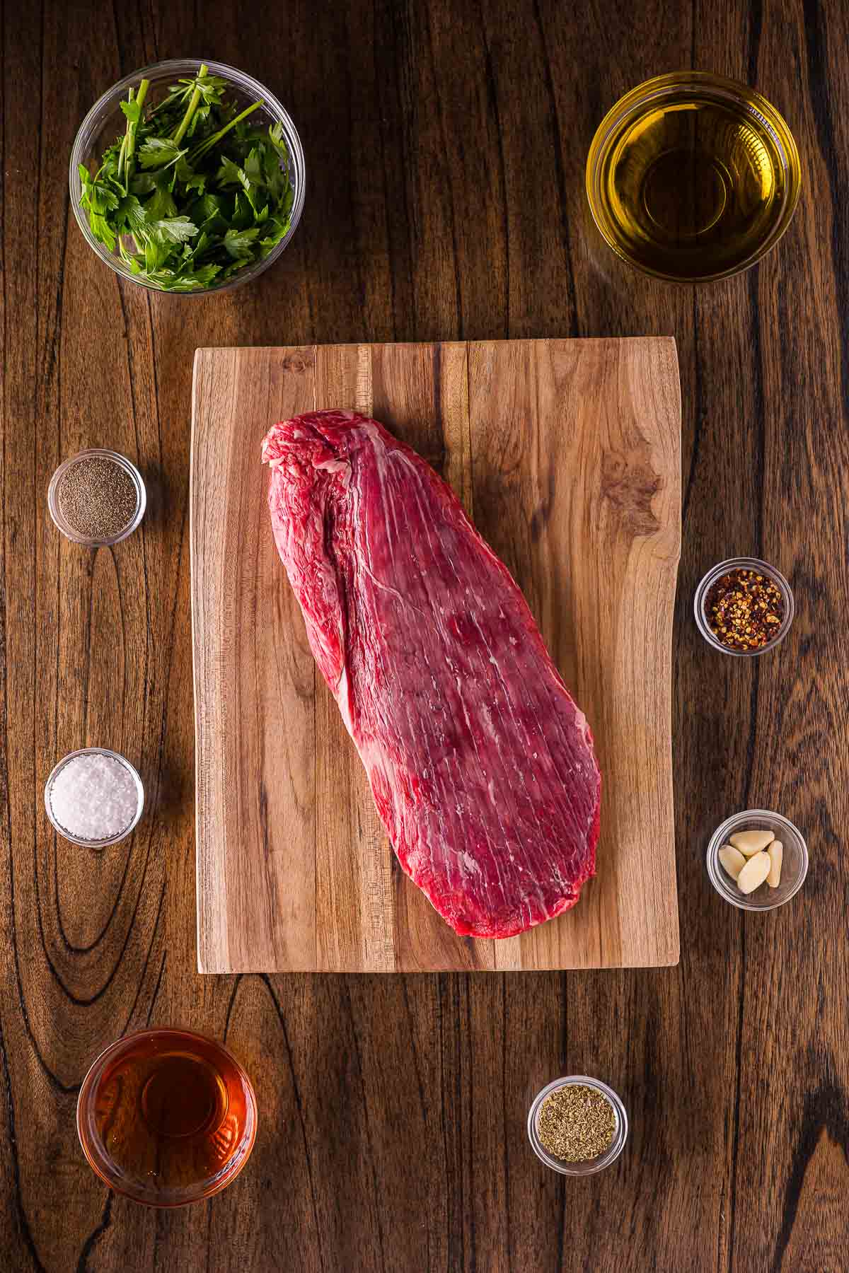 Raw flank steak on a wooden board surrounded by small bowls of parsley, olive oil, seasonings, garlic, and vinegar on a wooden surface.