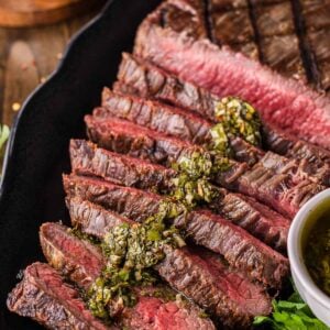 Sliced grilled flank steak served on a black plate with green herb chimichurri sauce on top, accompanied by parsley and a bowl of sauce.