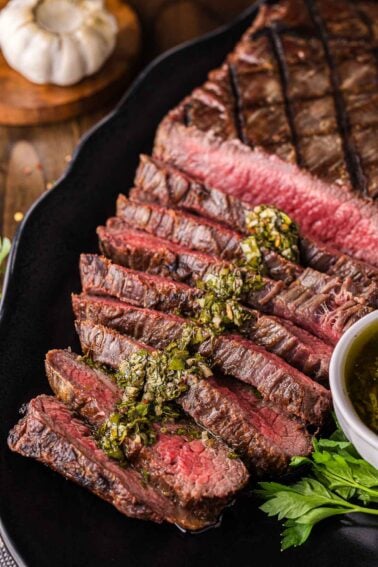 Sliced grilled flank steak served on a black plate with green herb chimichurri sauce on top, accompanied by parsley and a bowl of sauce.