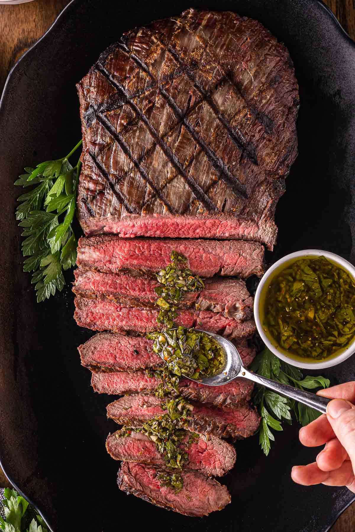 Grilled flank steak sliced and arranged on a black platter, topped with chimichurri sauce, with a small bowl of sauce and parsley garnish on the side.