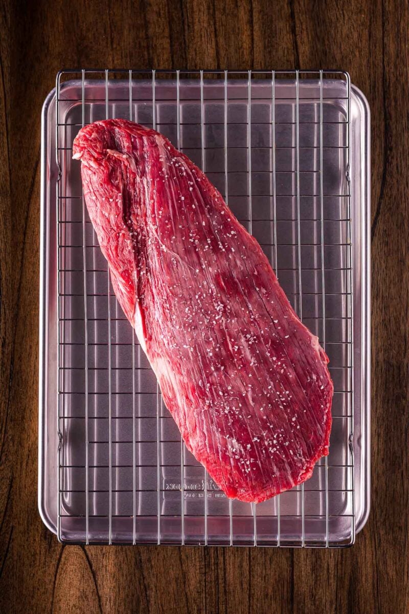 A raw, seasoned flank steak rests on a wire rack placed over a metal baking sheet on a wooden surface.