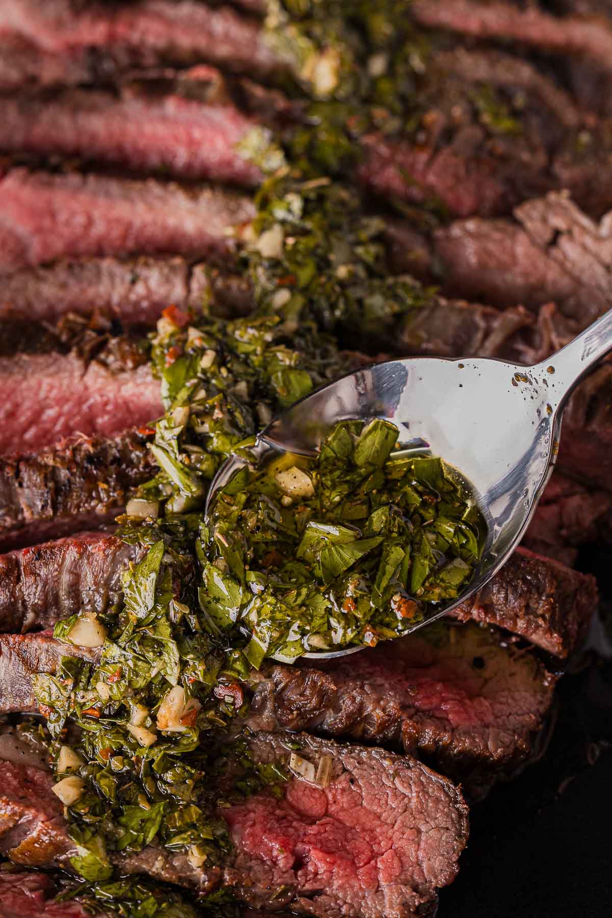 Sliced medium-rare steak topped with a green chimichurri sauce, with a spoon placed on top.