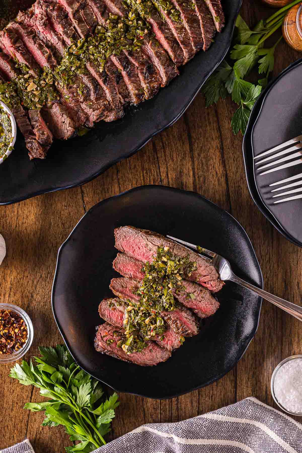 Sliced steak topped with chimichurri sauce is served on black plates, accompanied by herbs, seasoning, and utensils on a wooden table.