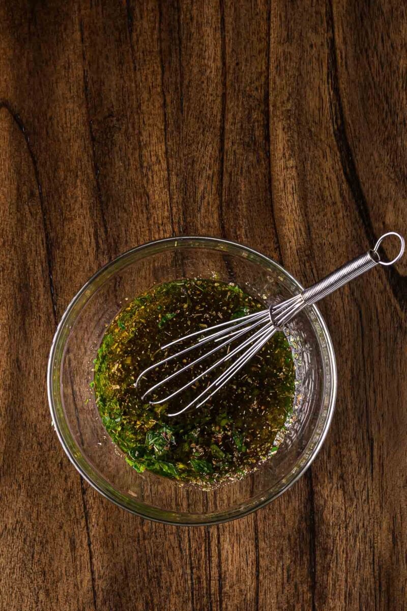 A glass bowl with a metal whisk contains a green herb chimichurri on a wooden surface.