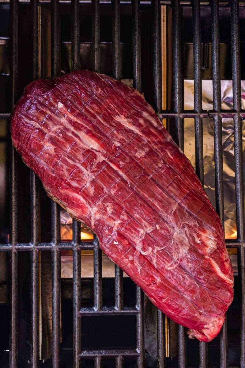 A raw flank steak with scored surface is placed on a grill above visible flames and metal grates.