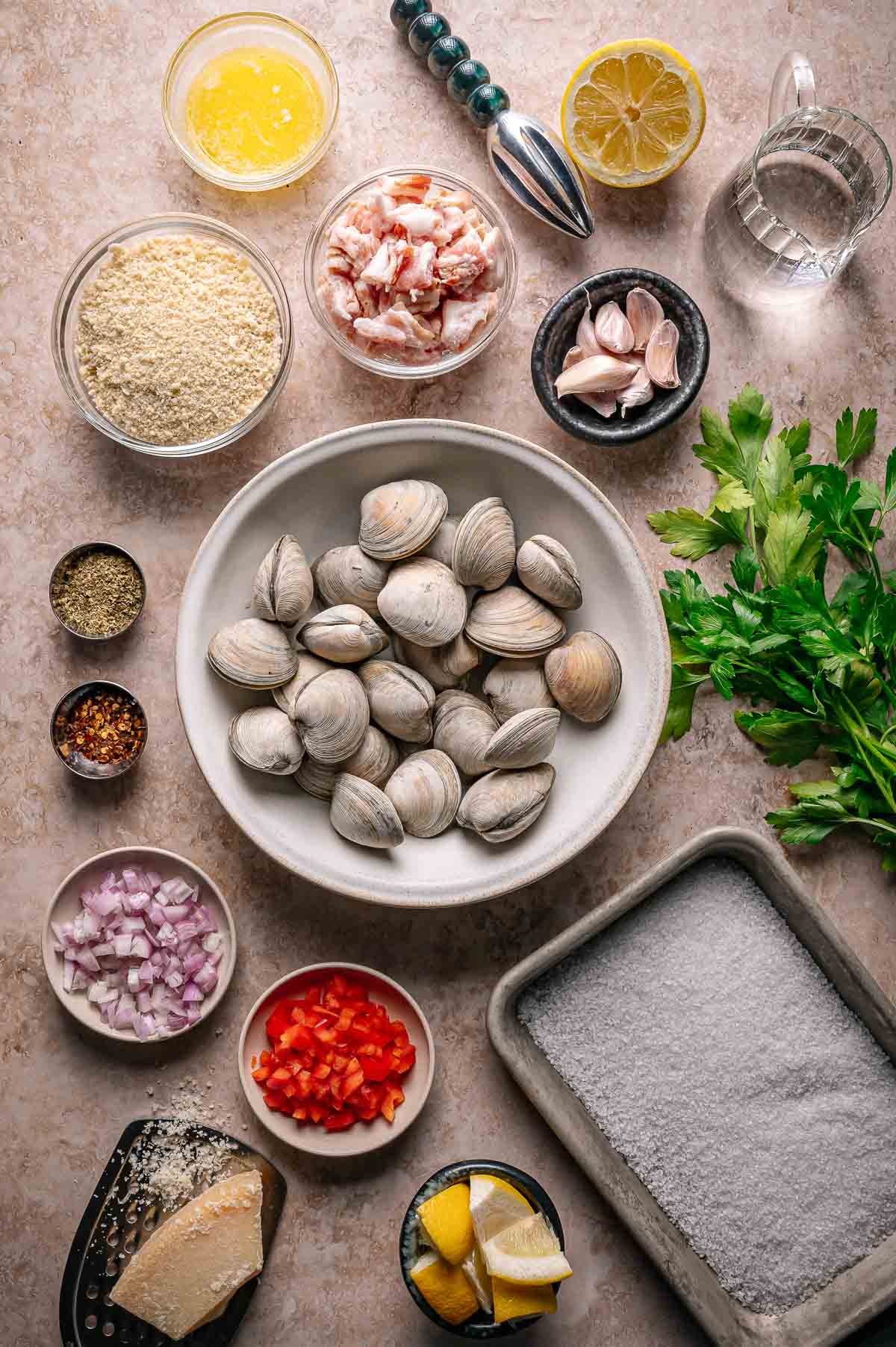 An overhead view of ingredients for stuffed clams casino, including clams, breadcrumbs, bacon, garlic, lemon, parsley, red pepper, shallots, grated cheese, butter, and seasonings on a countertop.
