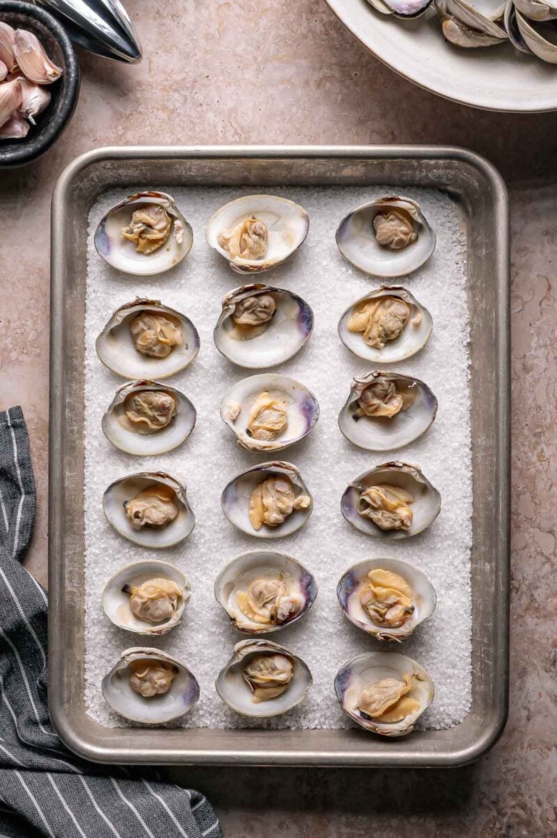 A baking tray with raw clams on the half shell arranged on a layer of coarse salt, ready for cooking. A striped kitchen towel and bowls are nearby.