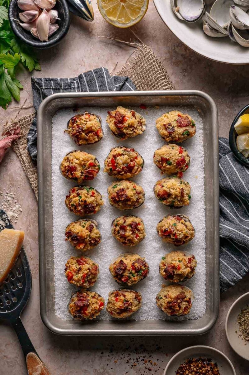 A baking tray with 18 stuffed clams casino on parchment paper, surrounded by ingredients like garlic, parsley, lemon, and grated cheese.