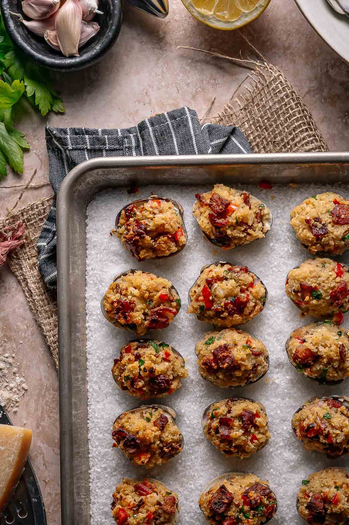A baking tray with stuffed clams casino filled with a breadcrumb and bacon mixture, arranged in neat rows on a bed of coarse salt.