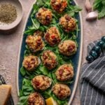 An oval platter with twelve stuffed clams casino on a bed of greens, garnished with lemon wedges, sits on a stone surface with herbs, cheese, garlic, and linen napkin nearby.
