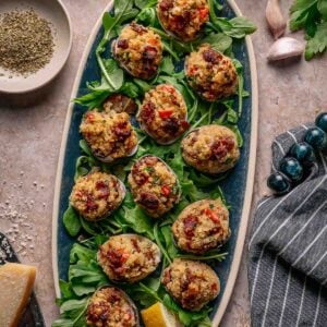 An oval platter with twelve stuffed clams casino on a bed of greens, garnished with lemon wedges, sits on a stone surface with herbs, cheese, garlic, and linen napkin nearby.