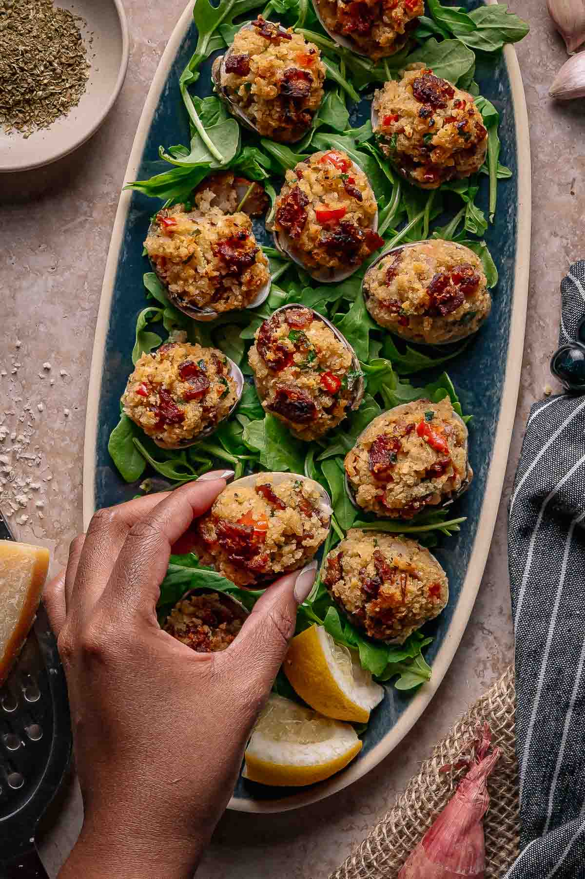 A hand picks up a stuffed clam from a platter arranged with greens, lemon wedges, and multiple baked stuffed clams casino.