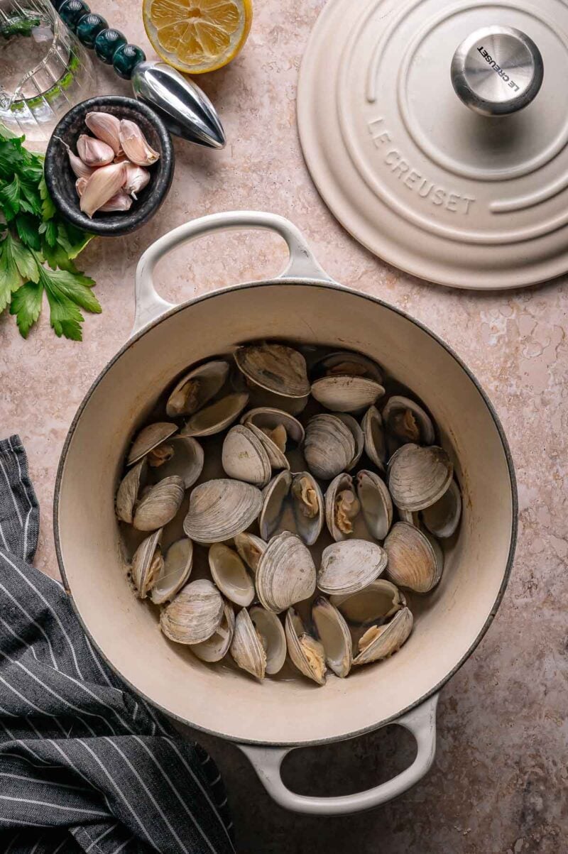 A white Dutch oven containing steamed clams sits on a countertop, surrounded by parsley, garlic, a lemon half, and a striped kitchen towel.