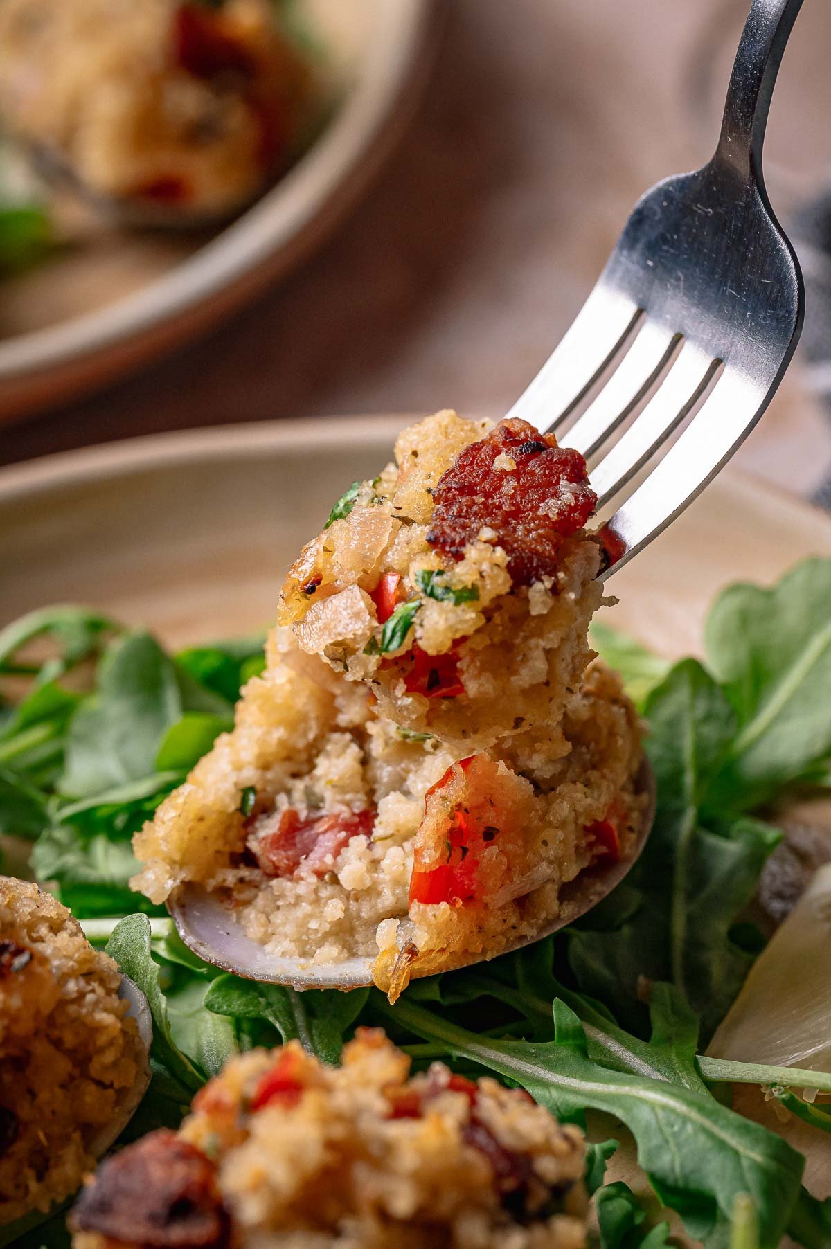 A fork holds a bite of stuffed breadcrumb mixture with bacon and herbs, taken from a clam casino resting on a bed of fresh arugula.