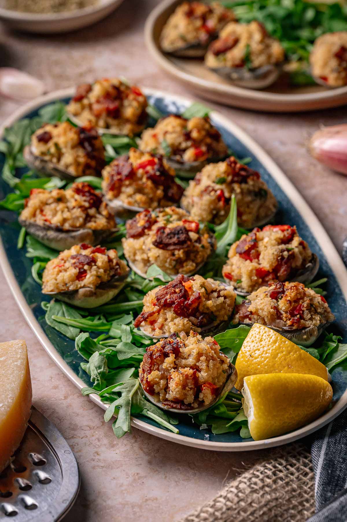 A platter of stuffed clams casino topped with breadcrumbs and herbs, served on a bed of arugula with lemon wedges on the side.