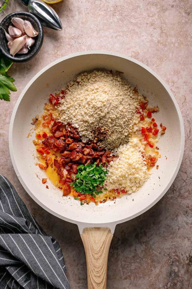 A frying pan on a countertop contains chopped cooked bacon, panko breadcrumbs, grated cheese, and chopped parsley arranged in separate sections.