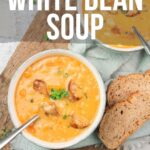 Two bowls of creamy white bean soup garnished with herbs and croutons, served with slices of bread on a wooden board.