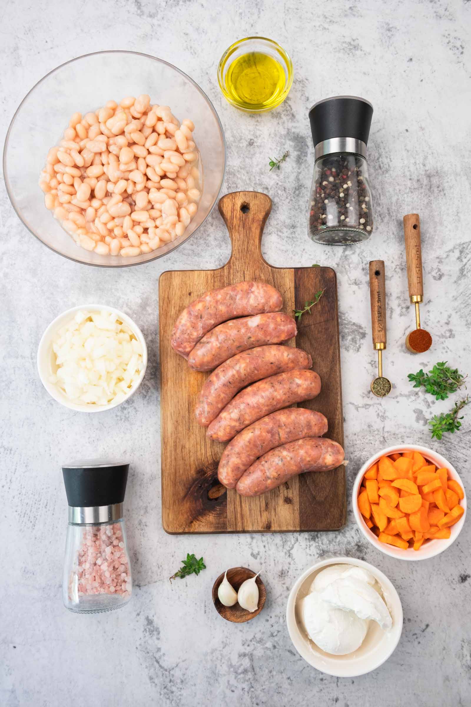 Top-down view of raw sausages on a cutting board, surrounded by bowls of white beans, chopped carrots, onions, garlic, spices, herbs, olive oil, and salt and pepper grinders.