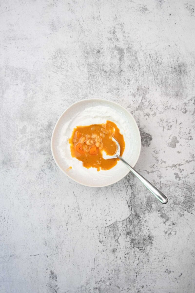 A white bowl containing sour cream with soup and white beans, with a spoon resting inside, on a light gray textured surface.