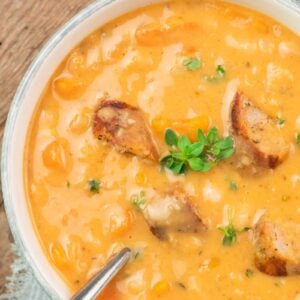 A bowl of creamy white bean soup with chunks of bread, sausage and a garnish of fresh herbs, served with a spoon on a light cloth.