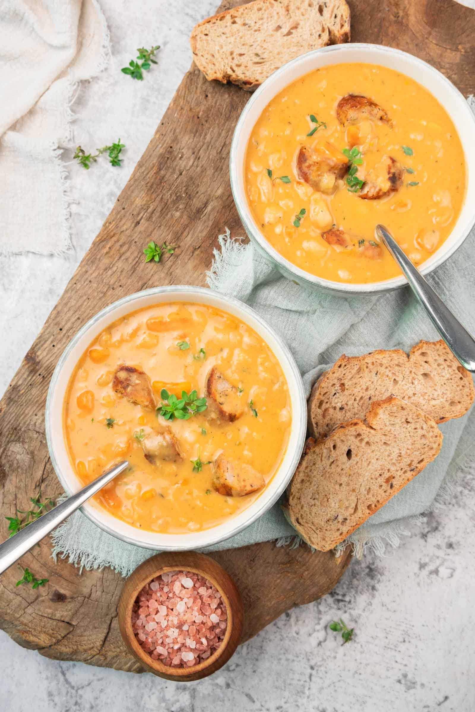 Two bowls of creamy white soup with sausage, garnished with herbs, served on a wooden board with slices of bread and a small bowl of pink salt.