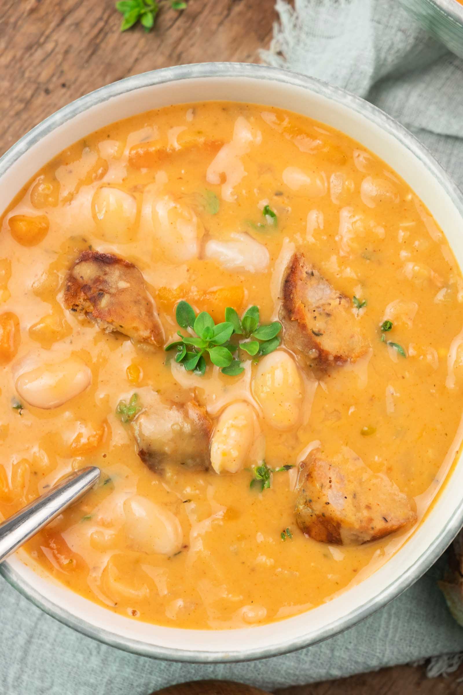A bowl of creamy white bean soup with sausage slices, garnished with fresh herbs, sits on a wooden surface with a spoon.