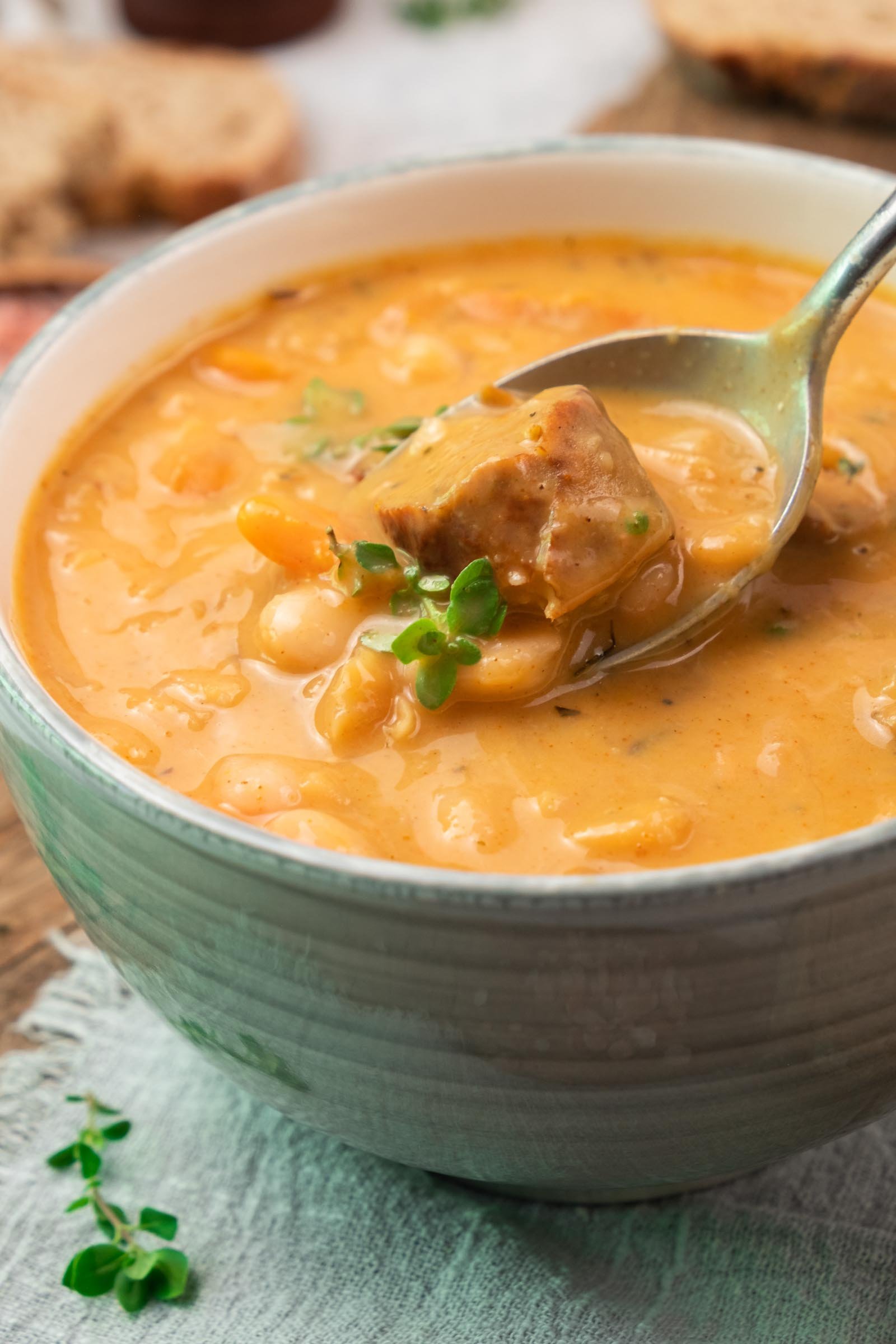 A close-up of a bowl of thick white bean soup with chunks of sausage, garnished with fresh herbs, and a spoon lifting a piece of meat from the soup.