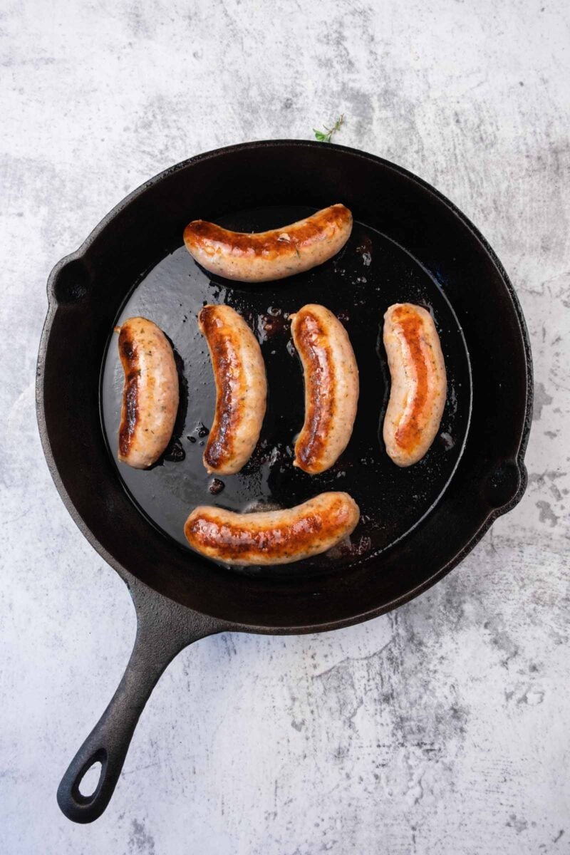 Six browned sausages are cooking in a black cast iron skillet on a light, textured surface.