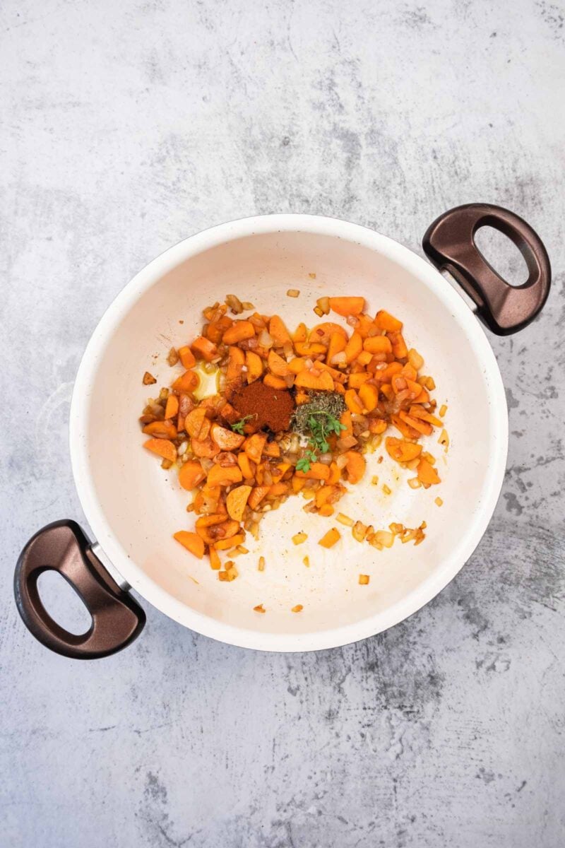 White pot with diced carrots, onions, and spices being sautéed on a light gray countertop.