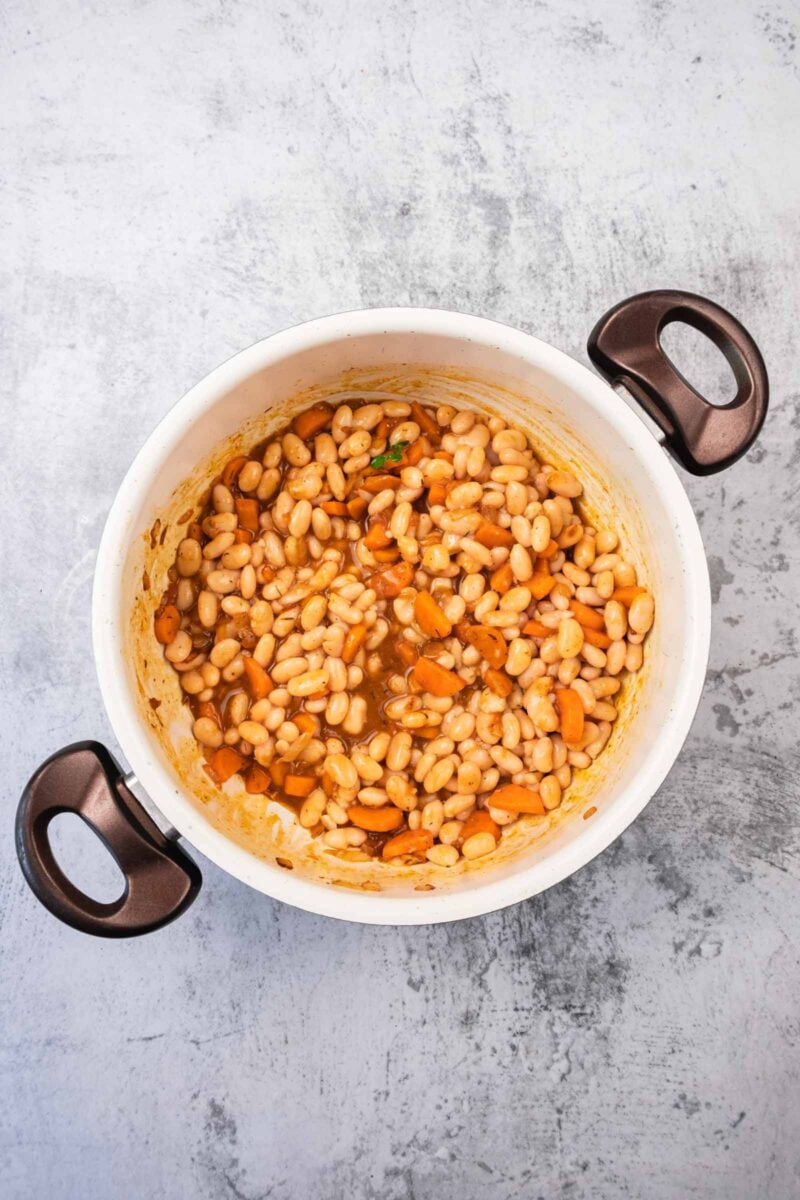 A white pot with black handles contains cooked white beans and sliced carrots in a light brown broth, set on a textured gray surface.