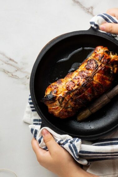 A person holds a cast iron pan with a cooked, tied roast on a striped towel. A brush, oil, and sauce are on the marble countertop.
