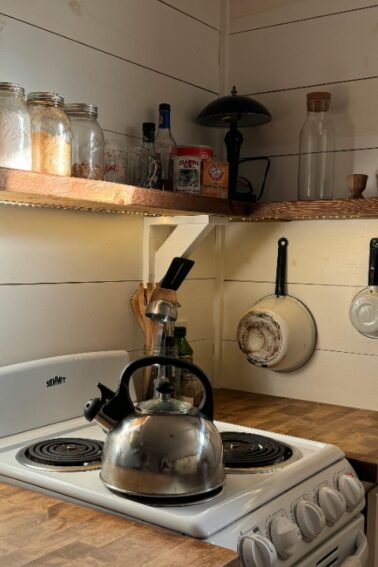 A small kitchen with a kettle on the stove, jars on open shelves, hanging pans on the wall, and a rolling pin on a wooden countertop. Sunlight streams in from the left.