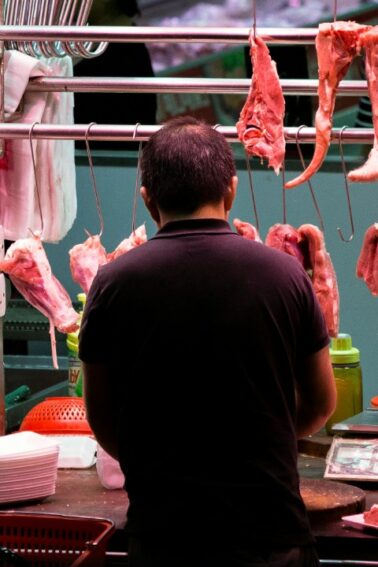 A person stands behind a counter in a butcher shop with raw meat hanging on hooks and placed on the counter.