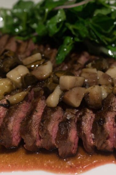 Sliced grilled steak with a mushroom topping, served with sauce and a side of fresh greens on a white plate.