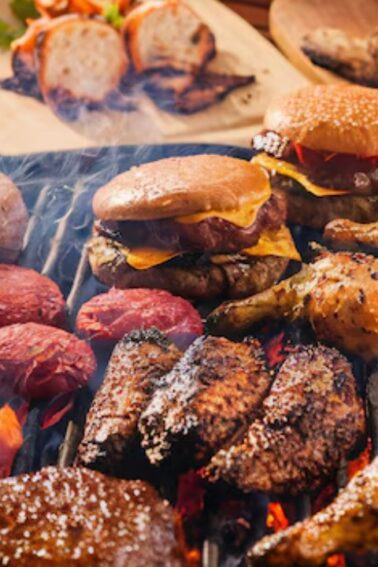 A variety of meats, burgers, and sausages are being grilled on a barbecue, with corn on the cob and side dishes visible in the background.