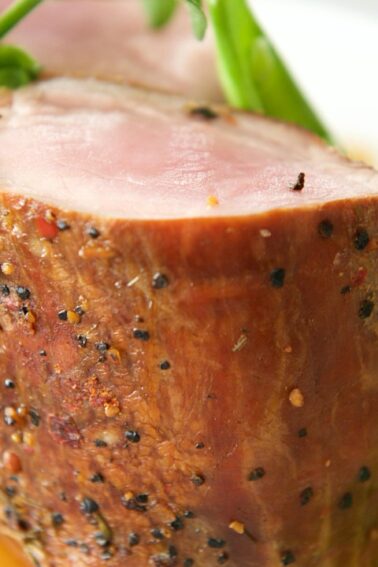 A close-up of a sliced, pepper-crusted pork tenderloin served with green pea puree and garnished with microgreens on a white plate.
