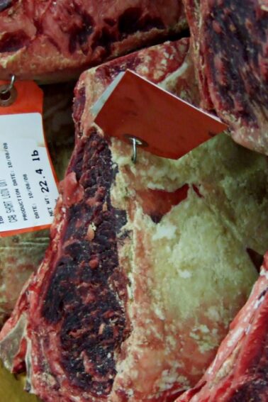 Several large cuts of raw beef, each tagged with labels indicating weight and details, are stacked side by side in a storage or aging facility.