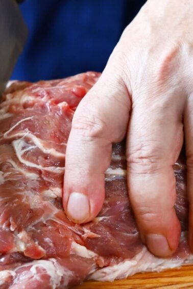A person slices a large piece of raw meat on a wooden cutting board with a knife.