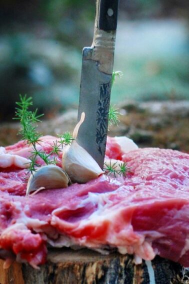 A raw steak with sprigs of herbs and garlic cloves on top is pierced by a knife, displayed outdoors on a piece of wood.