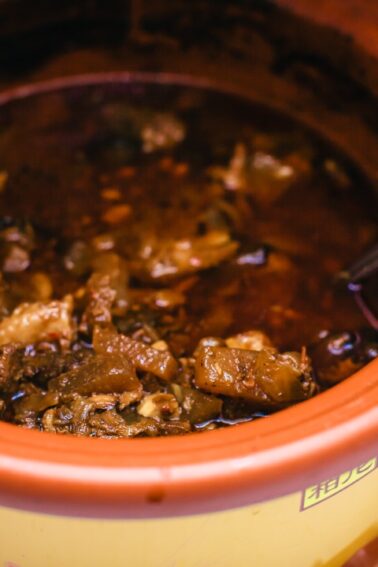 A brown ceramic pot filled with a dark, chunky stew or soup, with a metal spoon resting inside.