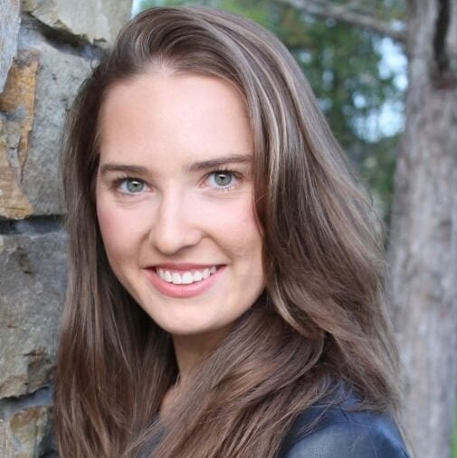 A woman with long brown hair and green eyes smiles while standing outdoors near a stone wall and trees.