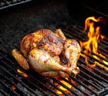 A whole seasoned chicken is grilling on an open flame barbecue grill, with visible flames and grill marks on the meat.