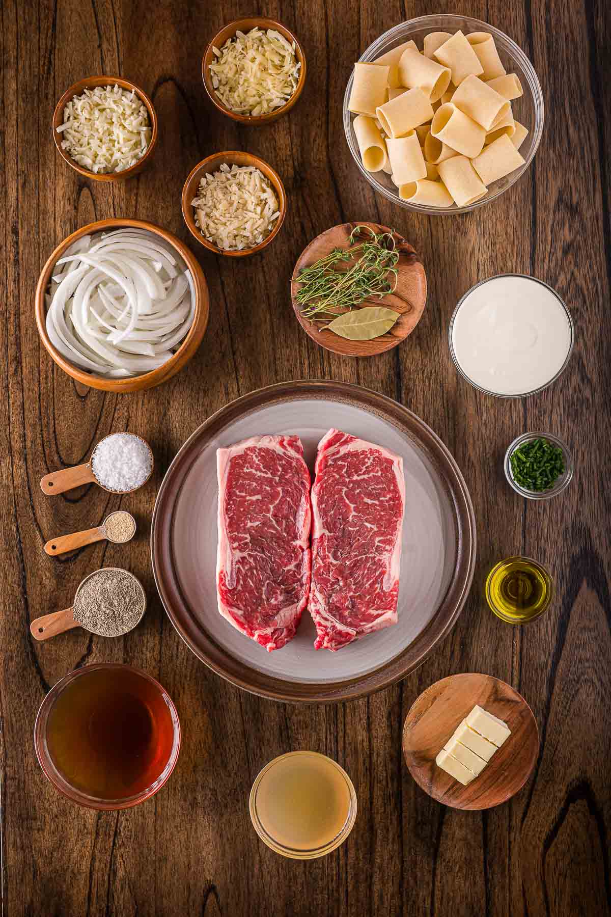 Overhead view of raw ribeye steaks, sliced onions, dry pasta, herbs, shredded cheeses, broth, cream, butter, olive oil, and seasonings arranged on a wooden surface.