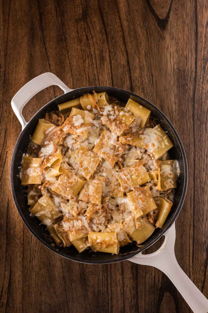 A white-handled skillet filled with french onion pasta topped with melted cheese sits on a wooden surface.