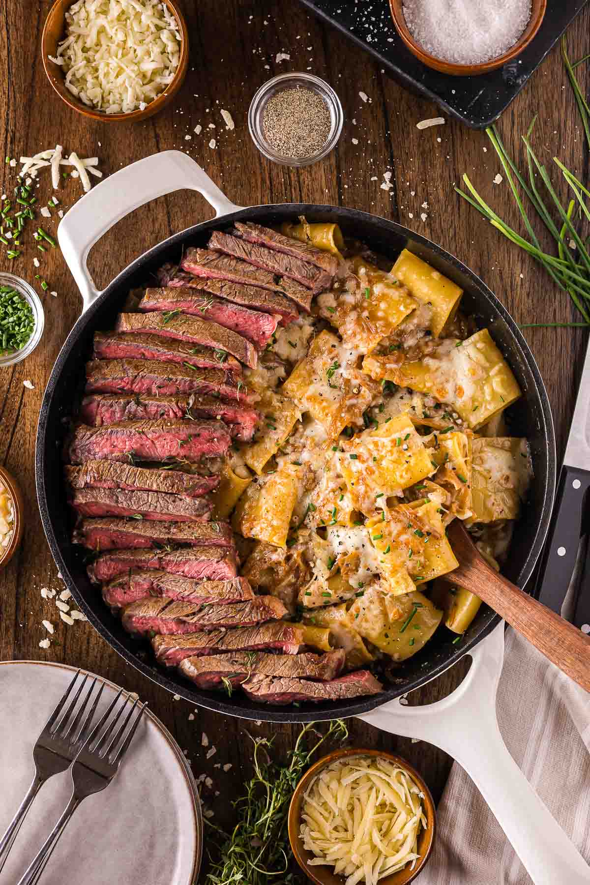 Sliced steak and creamy French onion pasta are served in a large skillet, garnished with herbs. Shredded cheese, seasonings, and fresh herbs are arranged around the skillet on a wooden table.