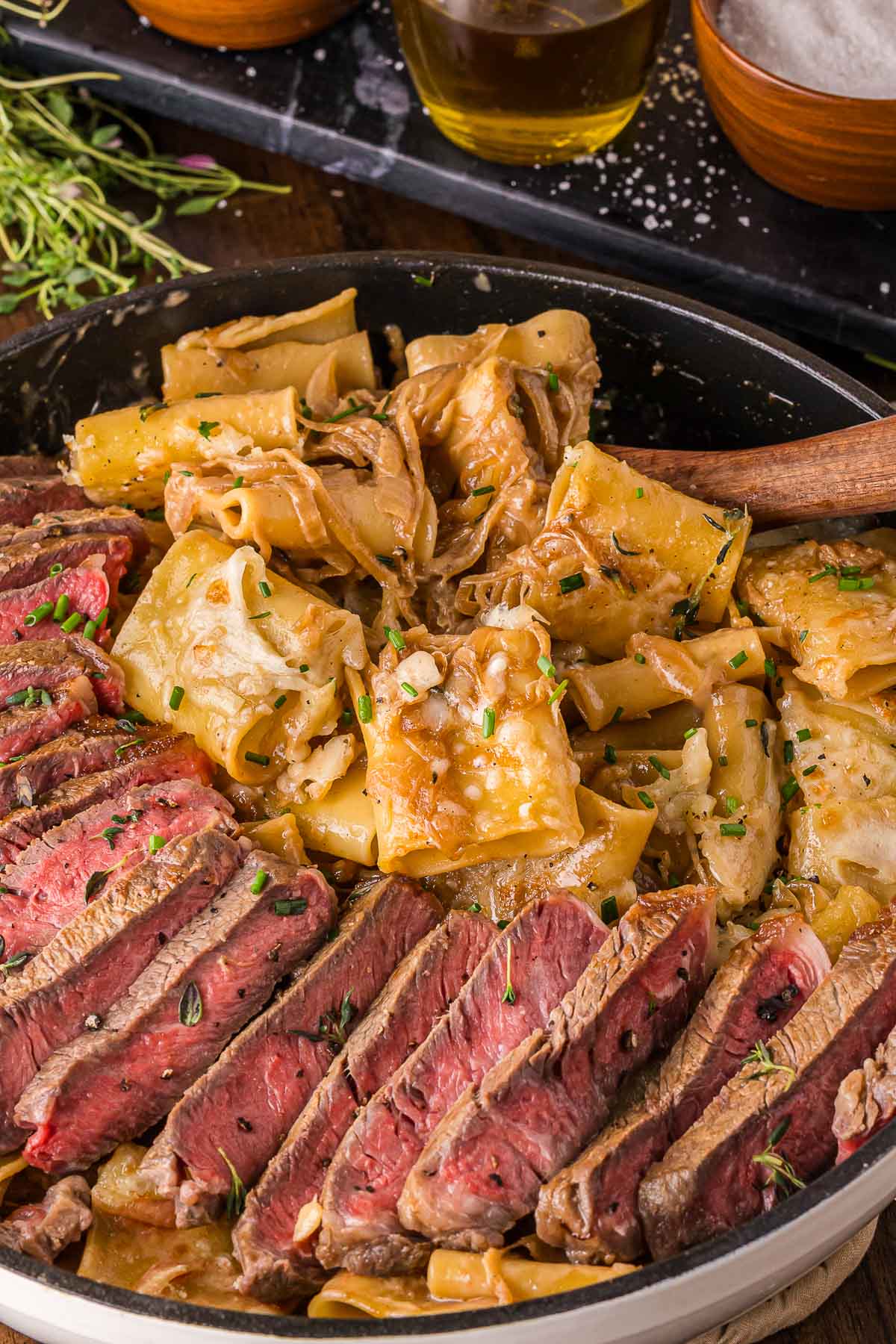 Sliced medium-rare steak arranged next to wide pasta with french onion sauce in a skillet, garnished with herbs.
