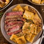 A plate with sliced steak and French onion pasta in a creamy onion sauce, garnished with herbs, next to a bowl of pasta and additional servings on the table.