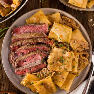 A plate with sliced steak and French onion pasta in a creamy onion sauce, garnished with herbs, next to a bowl of pasta and additional servings on the table.