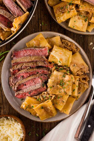 A plate with sliced steak and French onion pasta in a creamy onion sauce, garnished with herbs, next to a bowl of pasta and additional servings on the table.