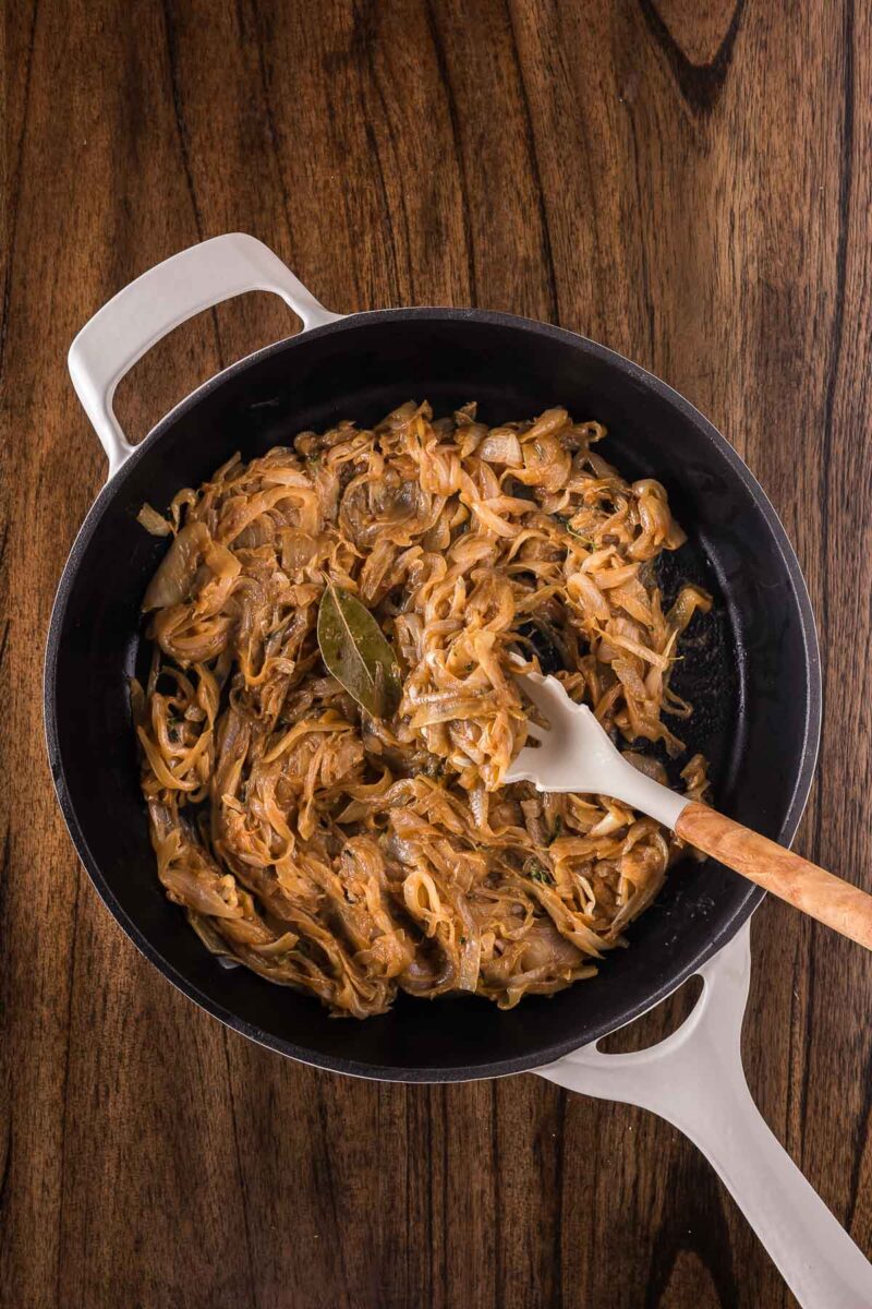 A white-handled pan on a wooden surface contains caramelized onions cooked with a bay leaf, being stirred with a spatula.